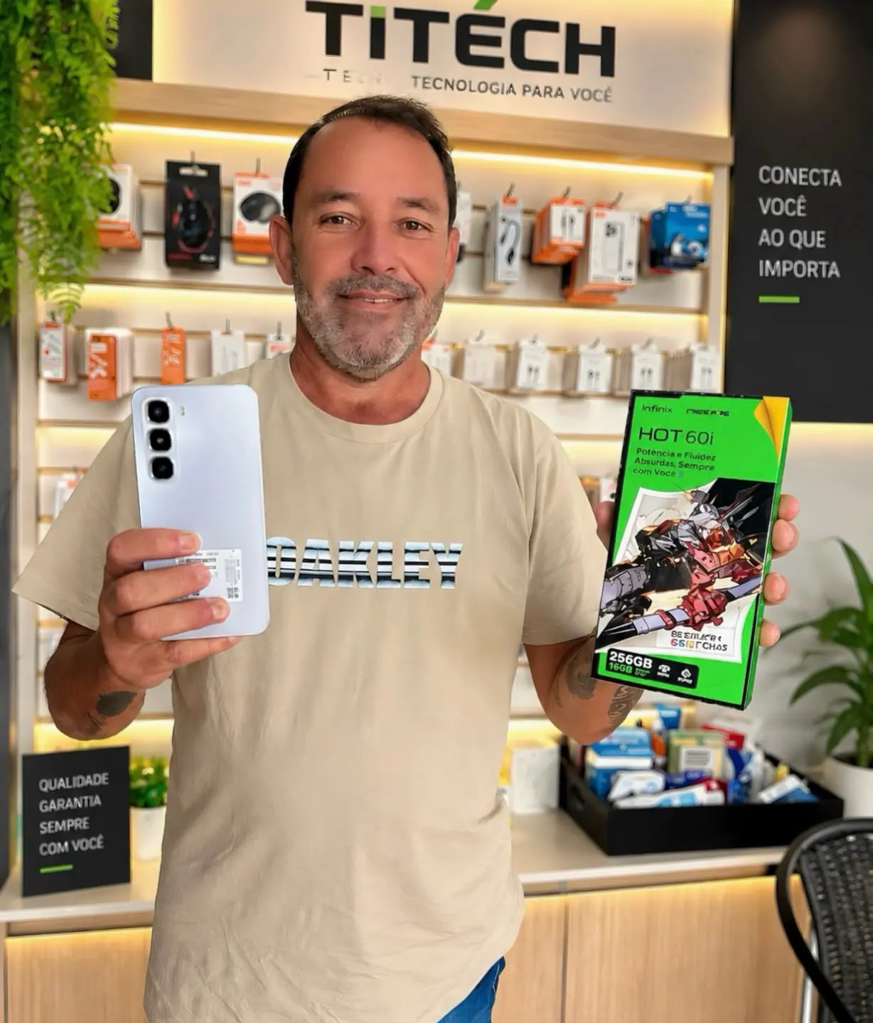Cliente segurando um celular e a caixa de um aparelho novo, na frente da parede de acessórios da loja